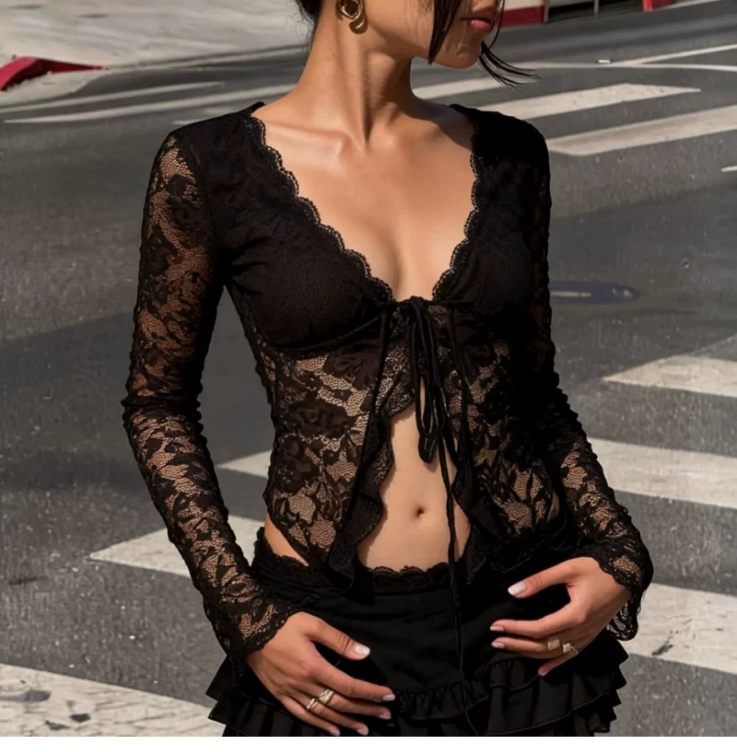 A woman wearing a black sheer lace long-sleeve top with a deep V-neckline, lace cups, and a front tie closure, photographed outdoors on a street crosswalk.
