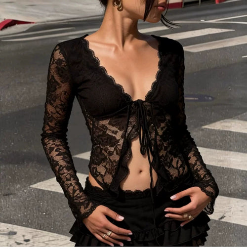 A woman wearing a black sheer lace long-sleeve top with a deep V-neckline, lace cups, and a front tie closure, photographed outdoors on a street crosswalk.