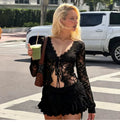 A woman walking outdoors on a street wearing a black lace long-sleeve top and matching black ruffled shorts, holding a green drink and carrying a brown shoulder bag.