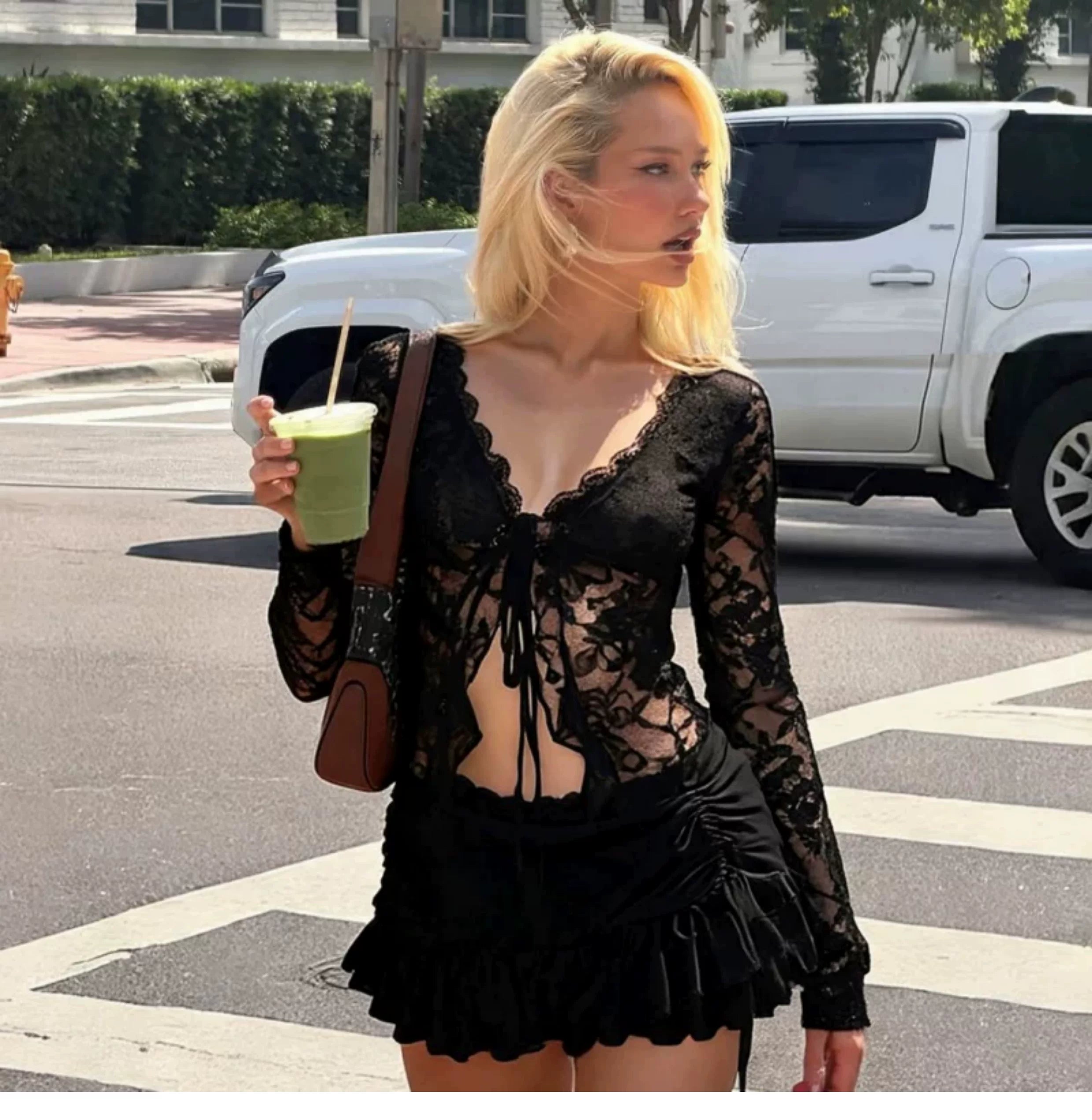 A woman walking outdoors on a street wearing a black lace long-sleeve top and matching black ruffled shorts, holding a green drink and carrying a brown shoulder bag.